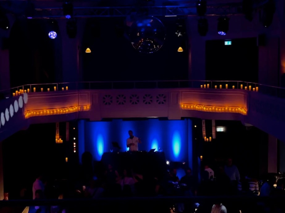 Blick von der Empore in einen Saal im blauen Licht, unten Phil am Pult vor der Tanzfläche, Kerzen an der Brüstung