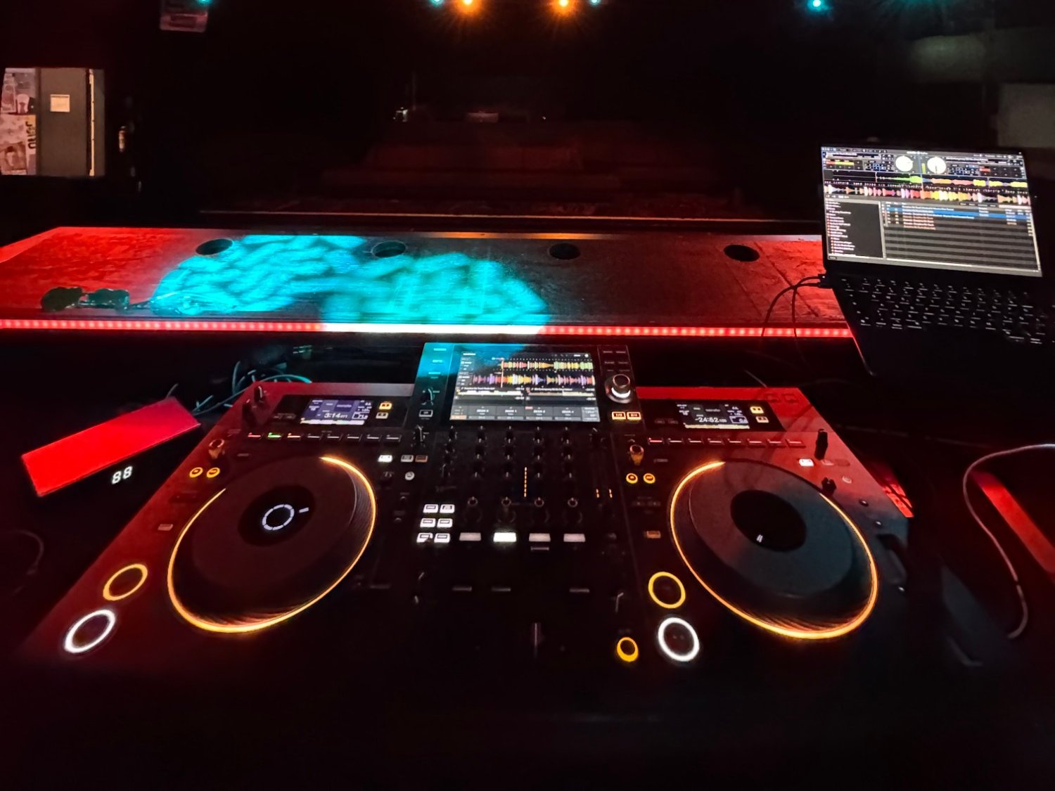 Aufgebautes Pult mit Laptop in einem Club, darüber Traversen in rotem und türkisem Licht