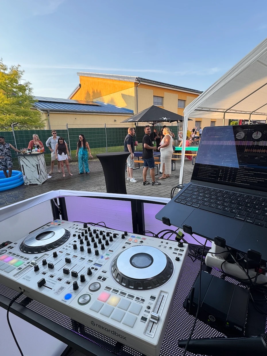 Blick vom DJ-Pult auf ein Sommerfest im Freien, Gäste an Stehtischen und Bierbänken unter blauem Himmel