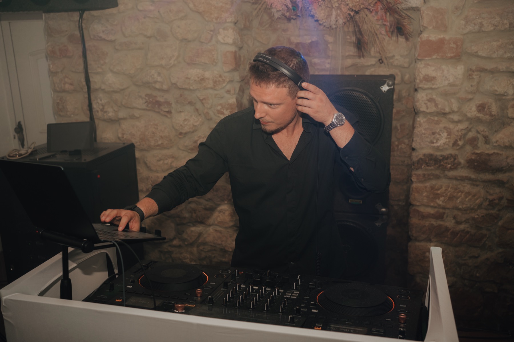 DJ mit Kopfhörern am Controller, hinter ihm eine Natursteinwand und ein Lautsprecher