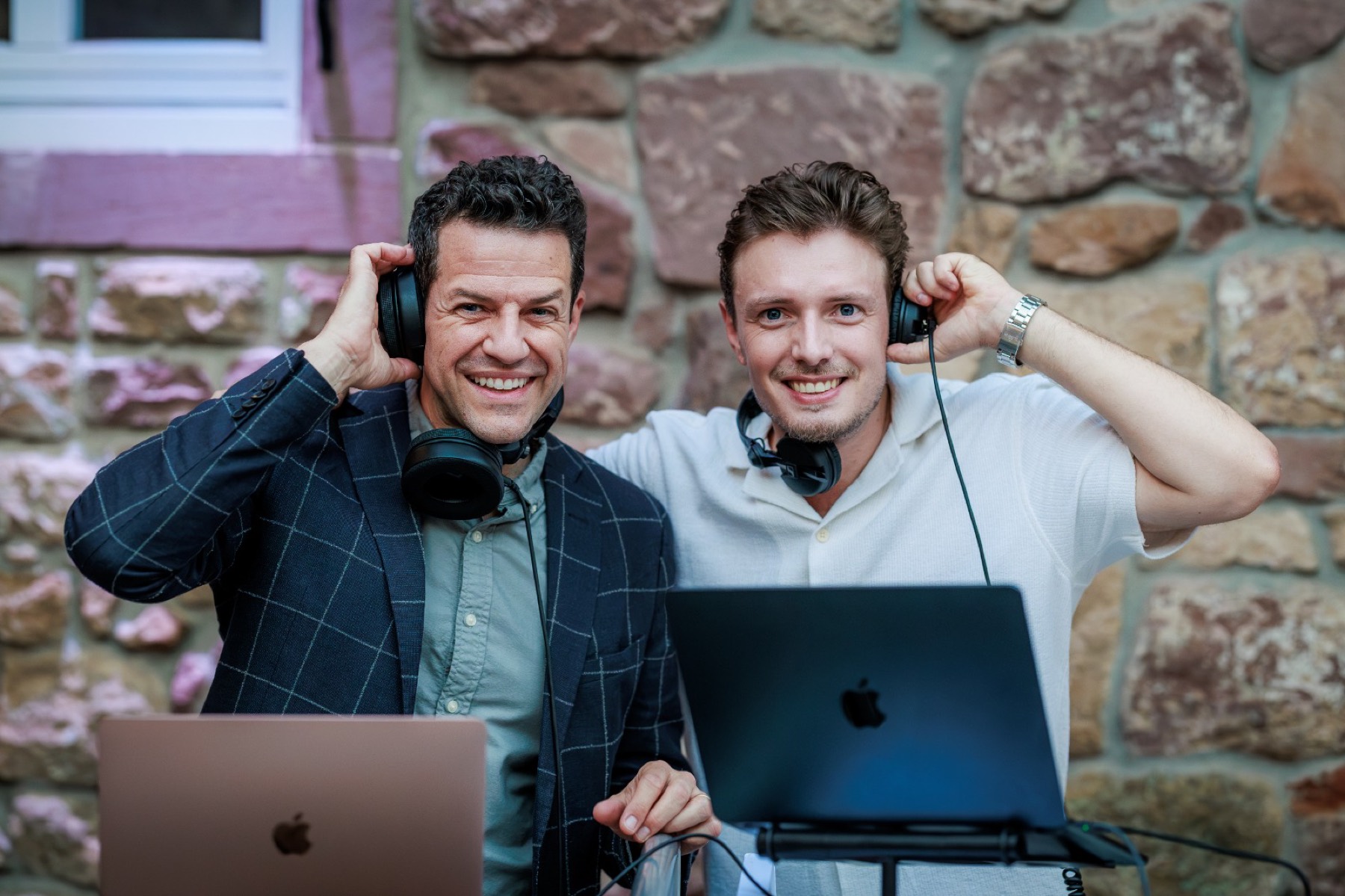 Zwei Männer mit Kopfhörern hinter ihren Laptops, hinter ihnen eine Natursteinwand