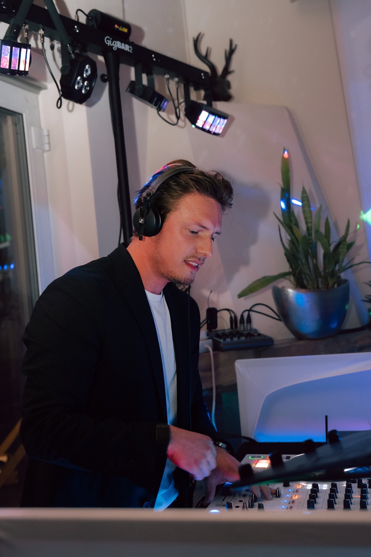 DJ im schwarzen Sakko mit Kopfhörern am Pult, über ihm eine Lichtleiste, daneben eine Pflanze